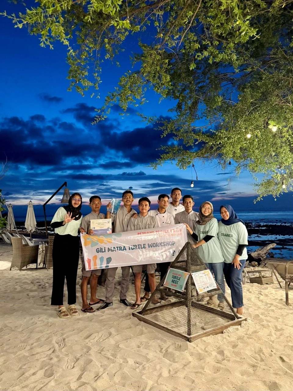 Group of people holding a banner on a beach during evening, with the ocean and sky in the background, and a turtle nesting area in the foreground.