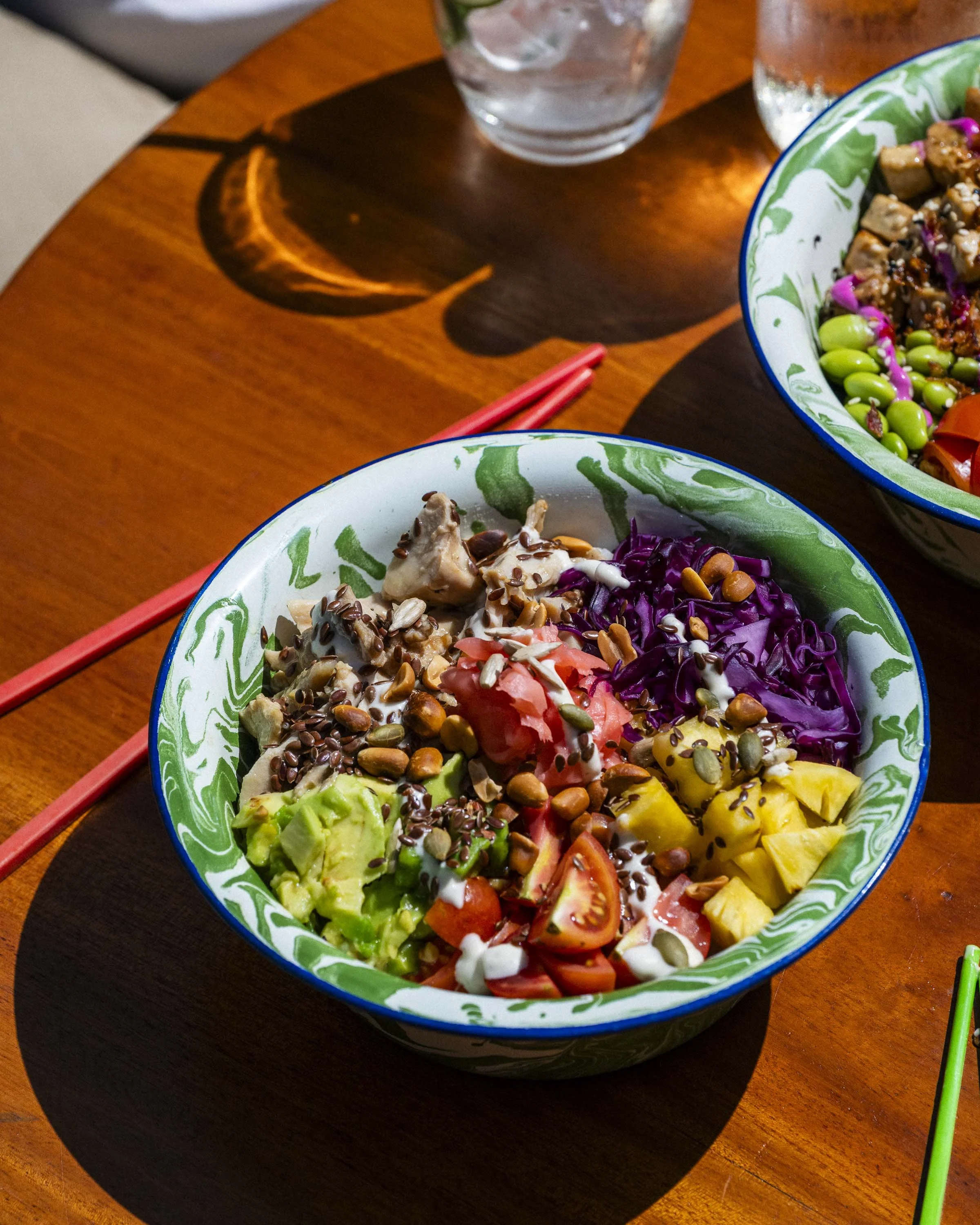 A colorful bowl of salad with tomatoes, avocado, purple cabbage, pineapple, and various seeds on a wooden table, with another bowl and glasses of water nearby.