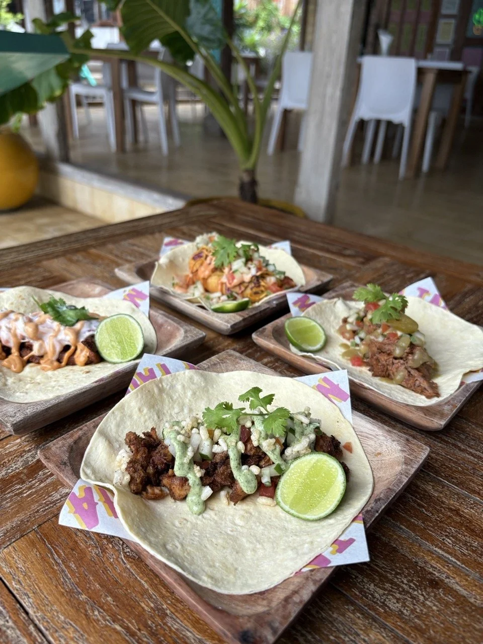 Four tacos on wooden plates garnished with lime wedges and cilantro, set on a wooden table in a restaurant.