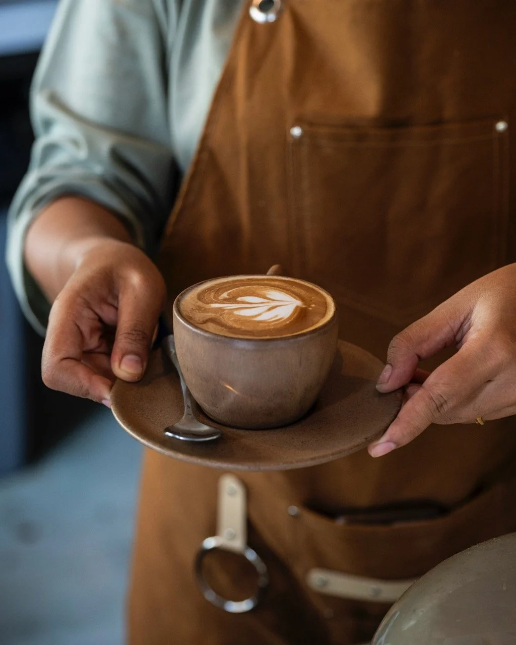 A person in a brown apron holds a ceramic cup of latte with latte art on top, sitting on a matching saucer in a coffee shop.