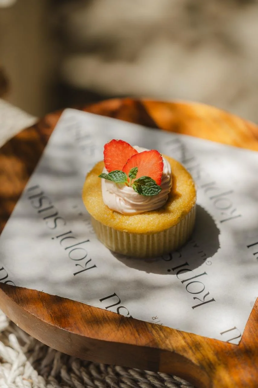 Cupcake topped with whipped cream, sliced strawberries, and mint leaves, placed on parchment paper inside a wooden bowl.