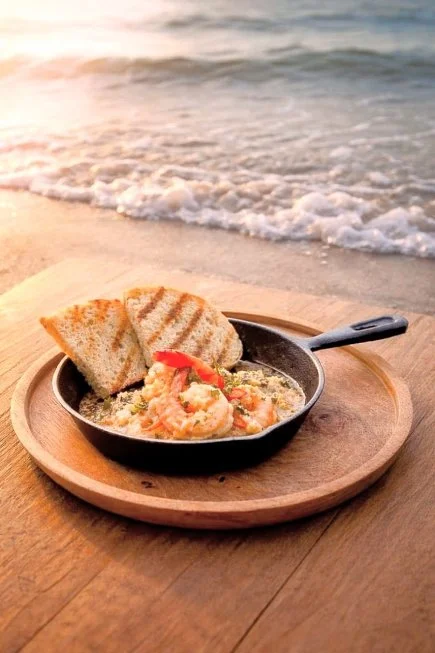 A skillet of shrimp, rice, and vegetables with two slices of toasted bread on a wooden tray at the beach, with ocean waves in the background during sunset.