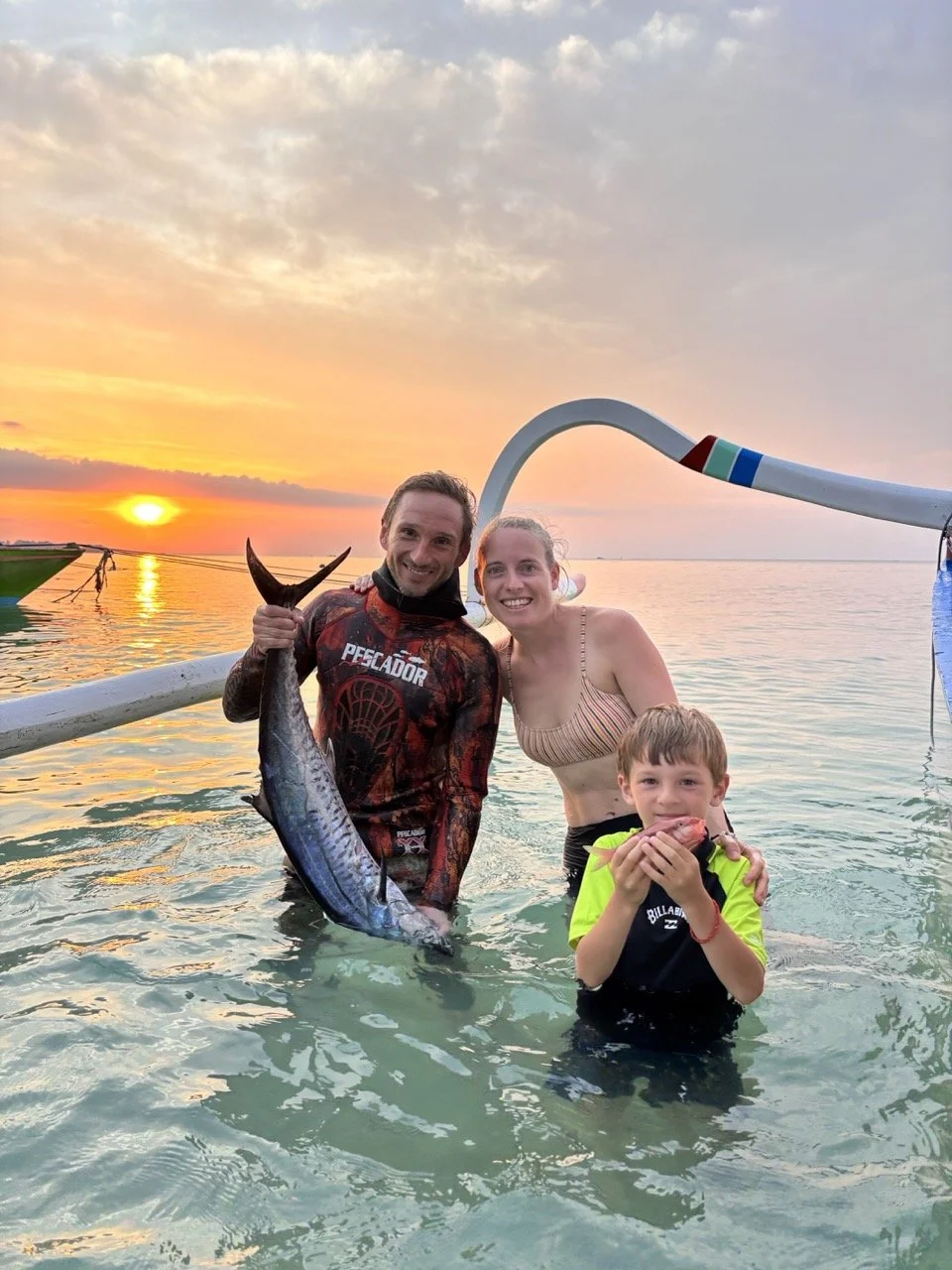 A family of three standing in the water at sunset, holding fish they caught during a beach outing. The man holds a large fish, and a boy holding a smaller fish. The woman is smiling with her arm around the man. The sky is partly cloudy with the sun setting on the horizon.
