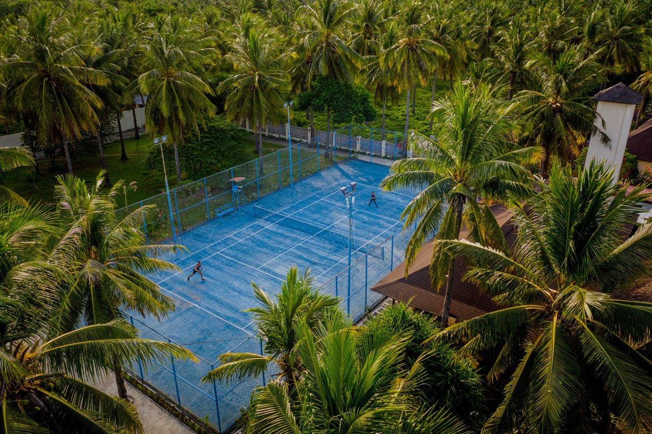 A blue outdoor tennis court surrounded by palm trees with two players on the court during daytime.