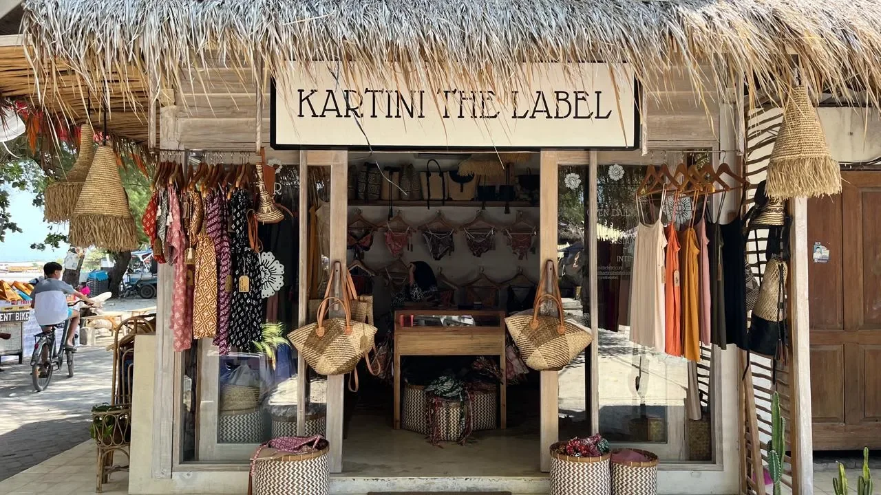A small beachside clothing and accessory shop with a thatched roof and wooden structure, displaying dresses, bags, and bathing suits.