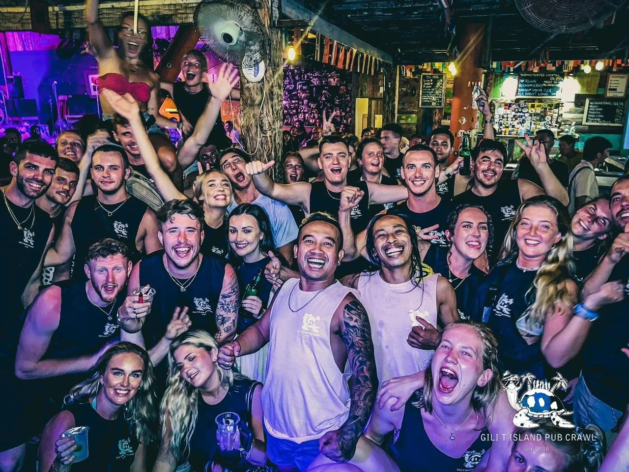 A large group of people at a lively Gili pub crawl party in a bar or club, smiling and celebrating, with vibrant lighting and wooden decor in the background.