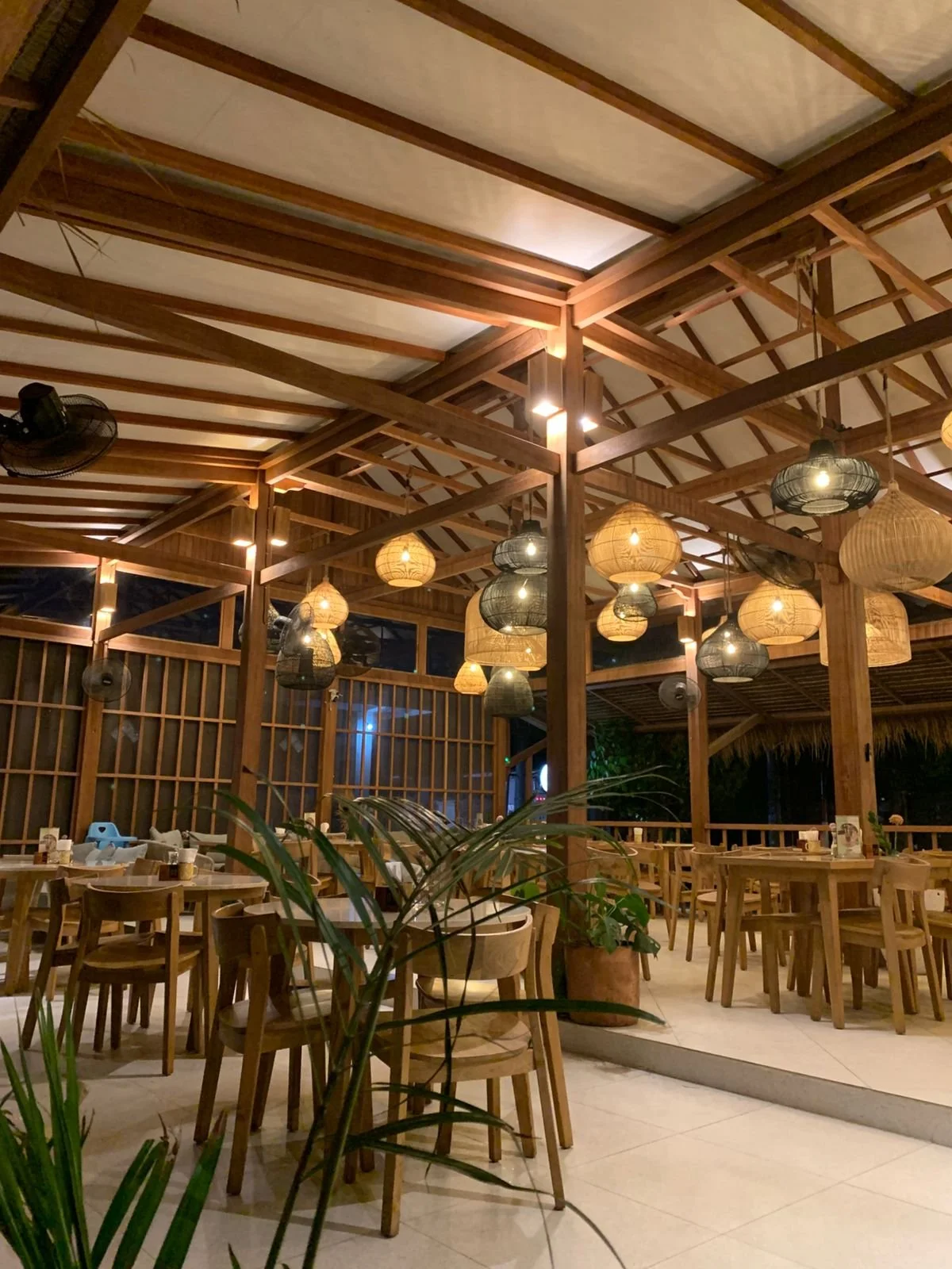 A beautifully designed open-air restaurant with wooden tables and chairs, hanging paper lanterns, and ceiling fans under a vaulted wooden ceiling with warm lighting.