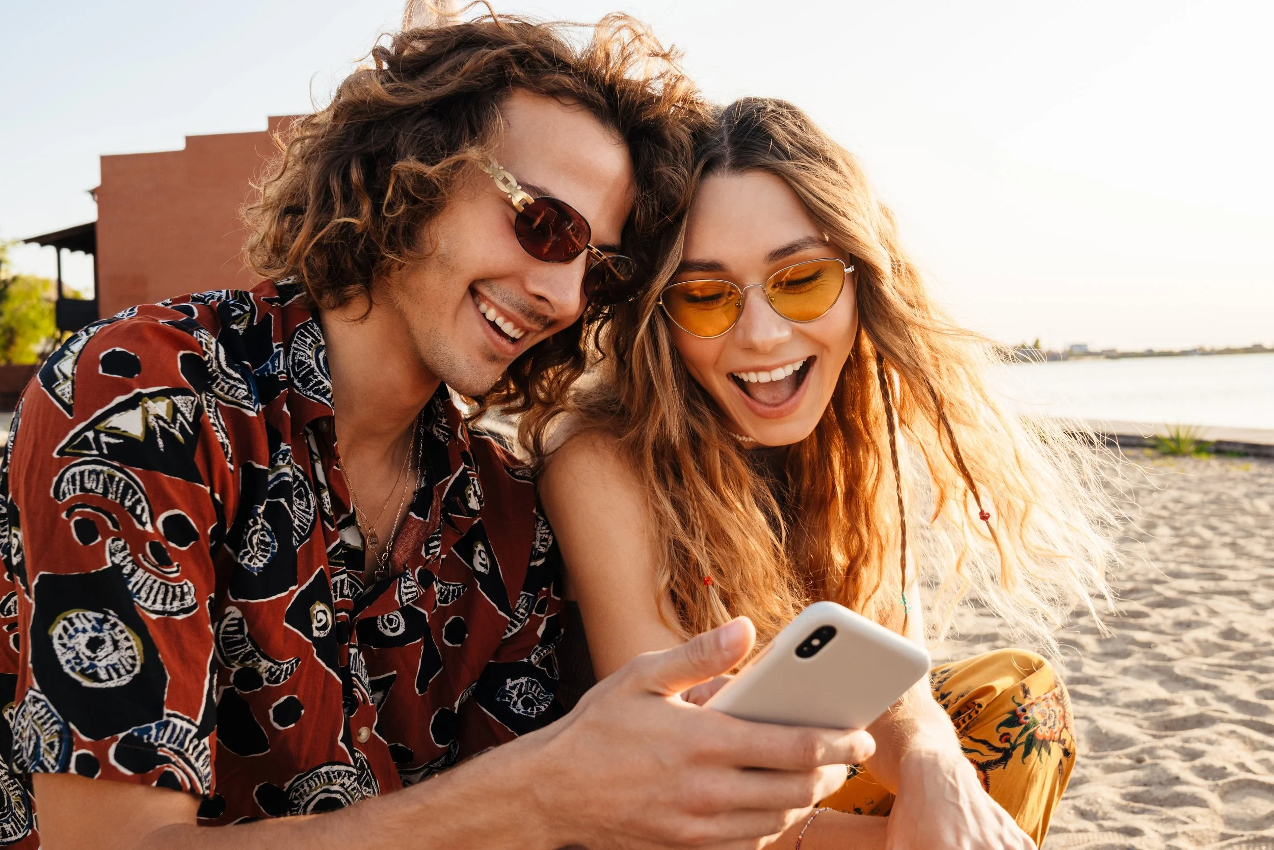 Woman with curly red hair wearing sunglasses and a striped tank top, lying on the ground next to a swimming pool, taking a selfie with her smartphone.