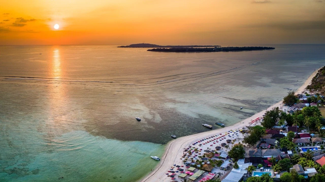 Aerial view of a beach at sunset with boats in the water, beach umbrellas, and a row of houses along the shoreline.