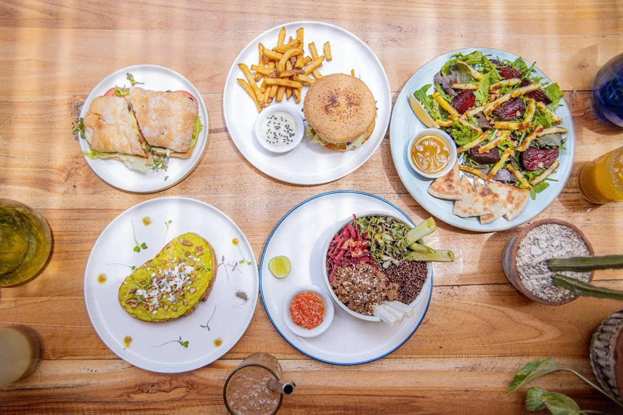 Assorted dishes on a wooden table, including a sandwich, burger, salad, avocado toast, and various beverages.