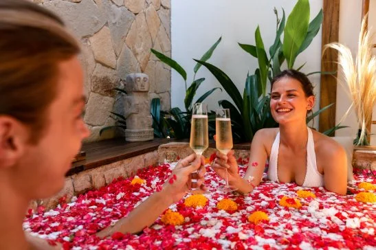 Two women in a flower-filled hot tub clinking champagne glasses, smiling, surrounded by green plants and stone decor.