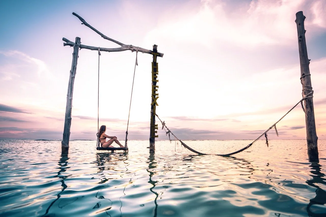 A woman sitting on a swing hanging from a wooden frame in the water at sunset, with a sign that reads 'THEE X L E' in the background.