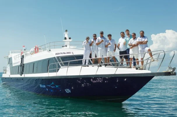 A group of nine people standing on the deck of a boat on the water, wearing white shirts and shorts, with a clear sky in the background.