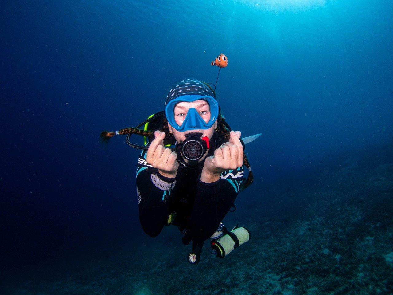 Woman scuba diving underwater, wearing blue mask and snorkel, holding a camera, with clownfish plush toy attached to gear.