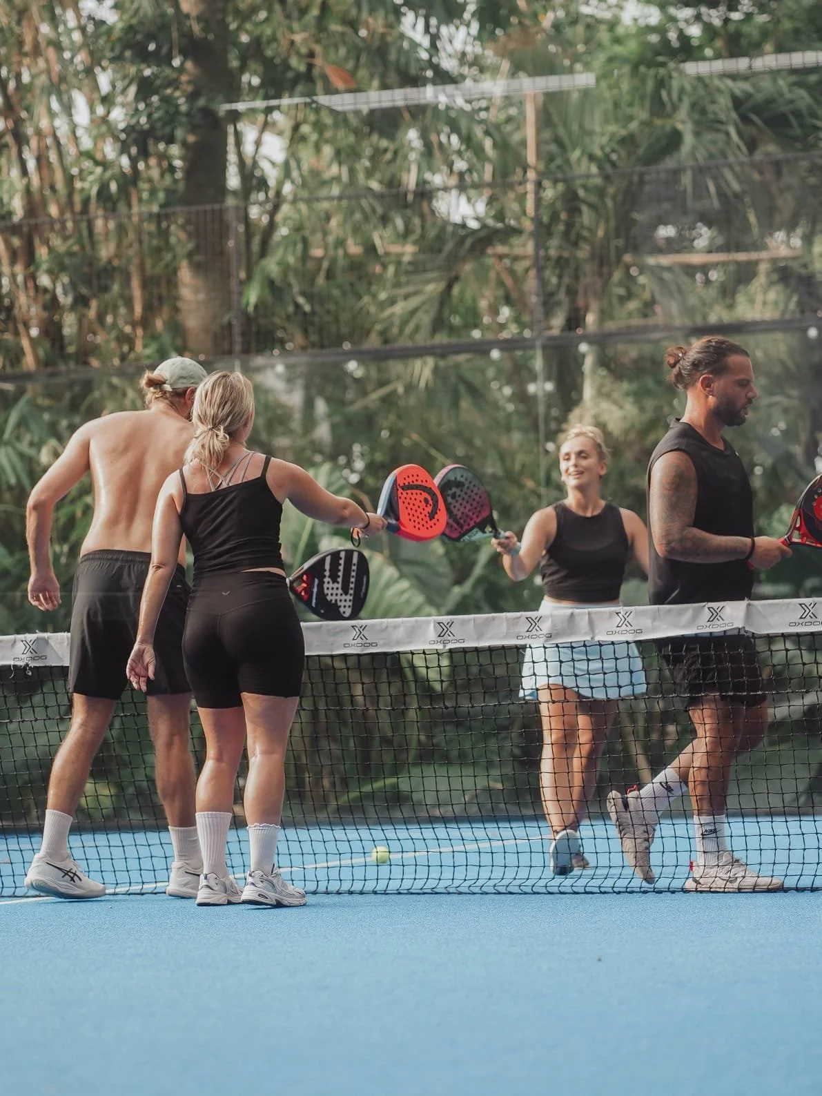 Group of four people playing pickleball on a court surrounded by trees, with a net in the foreground.
