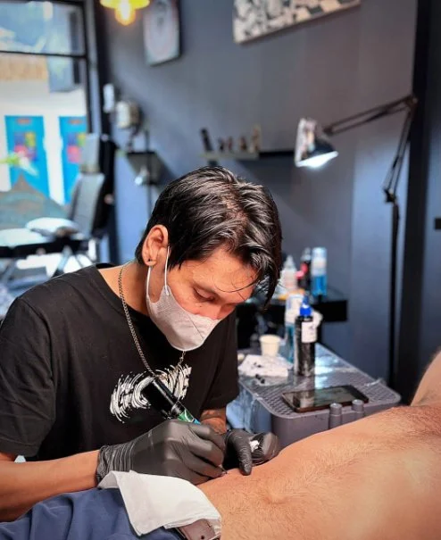 Tattoo artist wearing a face mask and gloves working on a client's arm in a tattoo studio.