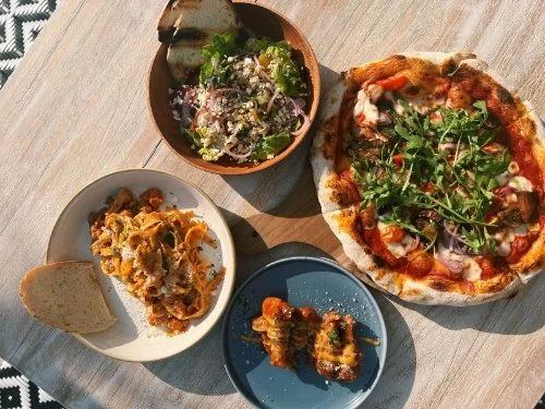 A meal consisting of a salad with greens, onions, and cheese in a wooden bowl, a slice of lasagna on a white plate with a piece of bread, a small pizza topped with arugula and vegetables, and two pieces of dessert, possibly cannoli, on a blue plate.