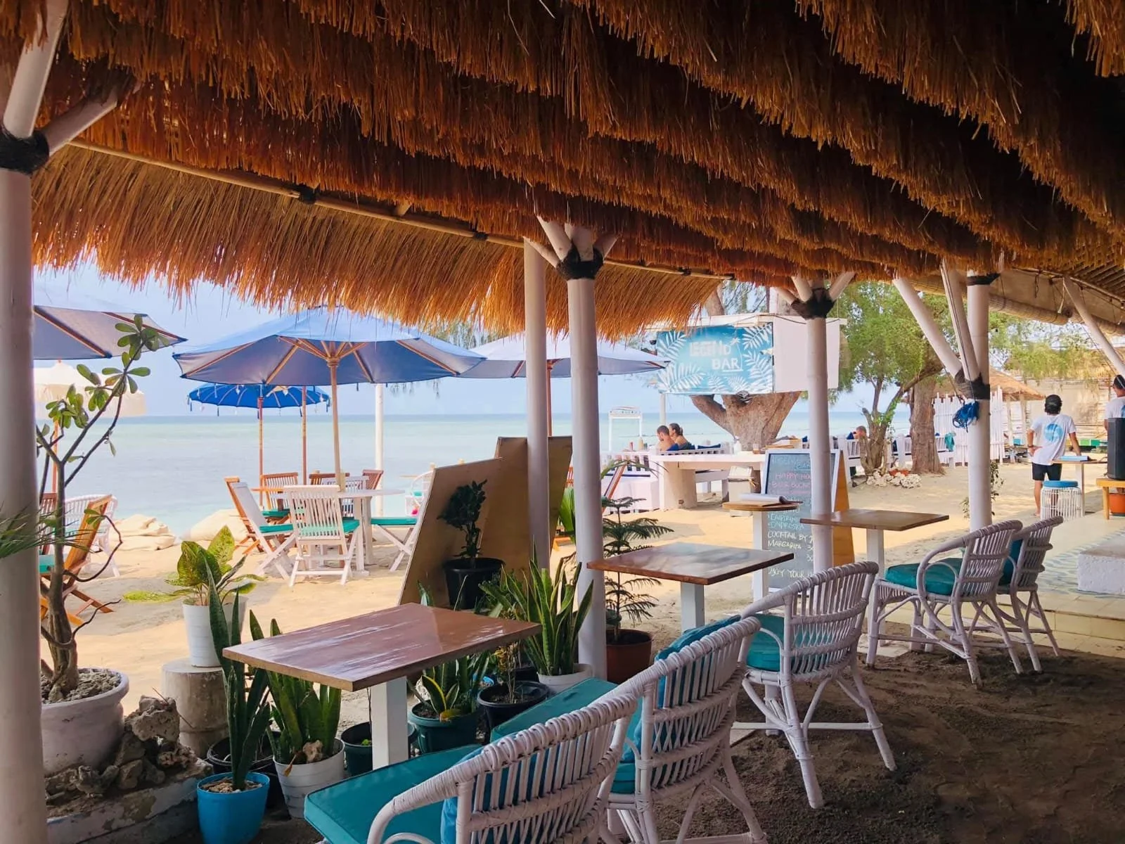 Beachside restaurant with wooden tables, white chairs with teal cushions, potted plants, and a thatched roof, overlooking the ocean with umbrellas and people in the background.