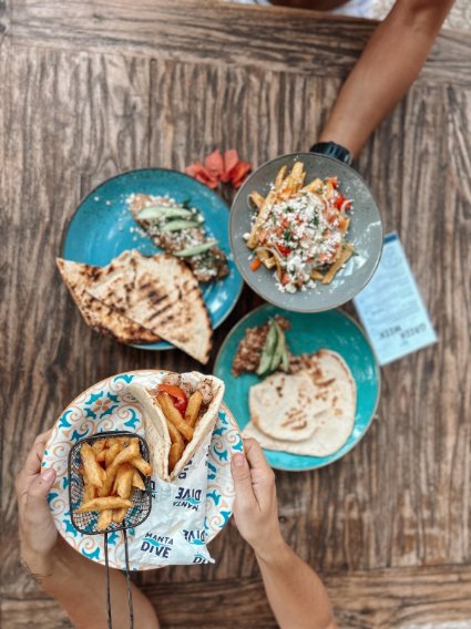 Overhead view of a wooden table with three plates of food and a hand holding a basket of fries in a pita. The plates include a salad, grilled chicken with rice, and a sandwich bun, with an arm reaching for the salad.