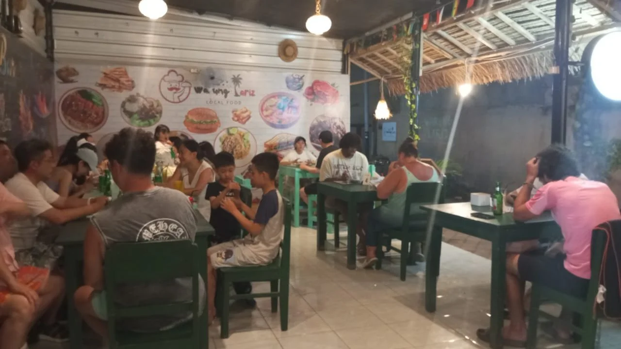 Interior of a restaurant with multiple tables filled with people, some eating and some using their phones, wall decorations featuring food images, and an open kitchen area.