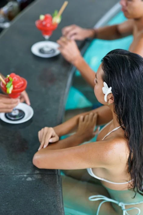 Two people in a pool sitting at a bar counter, each with a colorful frozen cocktail drink garnished with fruit and a cherry.