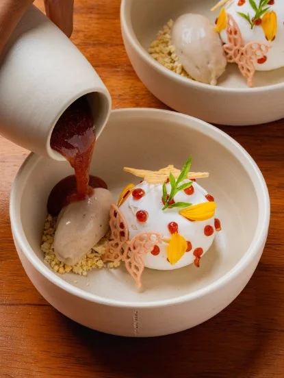 Pouring chocolate sauce over a dessert with white icing, red and yellow decorations, and a scoop of ice cream in a white bowl.
