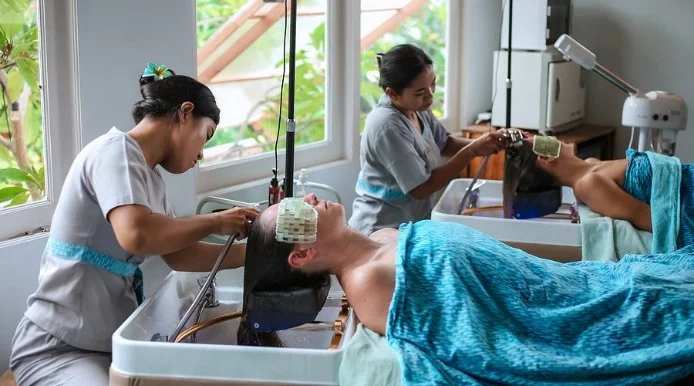 Two women are giving hair treatments to two clients in a hair salon, with both clients lying back with their heads in sinks.