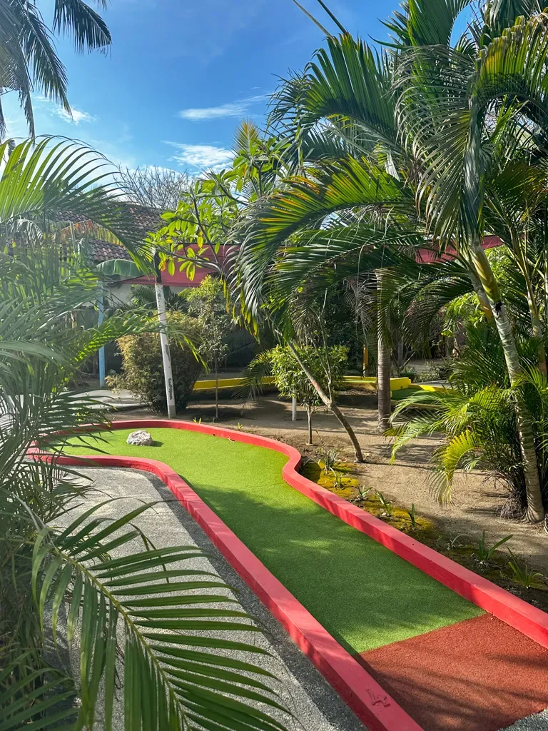 Miniature golf course with artificial turf, surrounded by tropical plants and palm trees, under a partly cloudy blue sky.