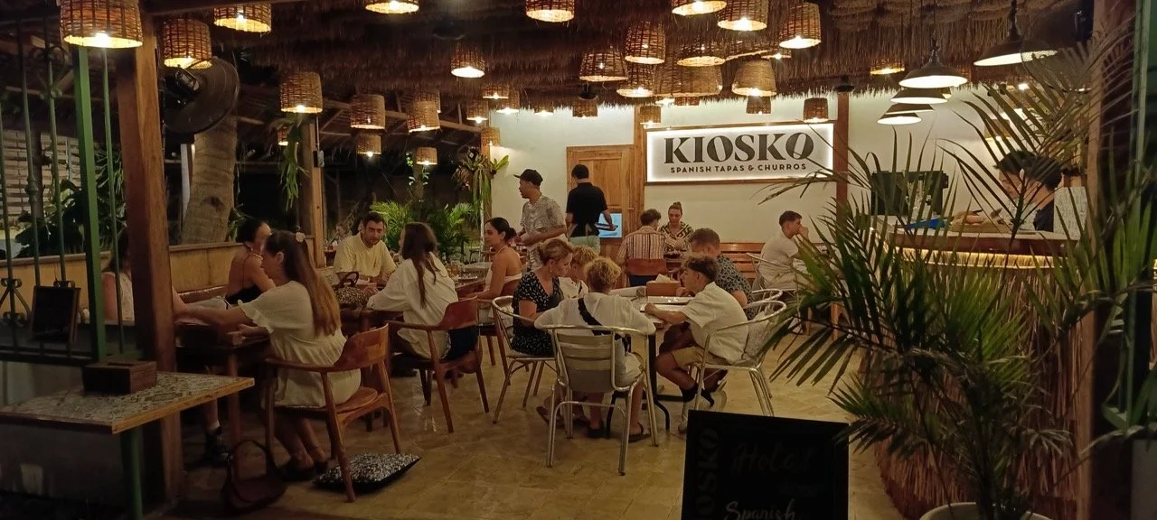 Inside a lively Spanish tapas and churrros restaurant, decorated with hanging wicker lamps, surrounded by lush green plants, with customers dining at various tables and a view of the kitchen area.