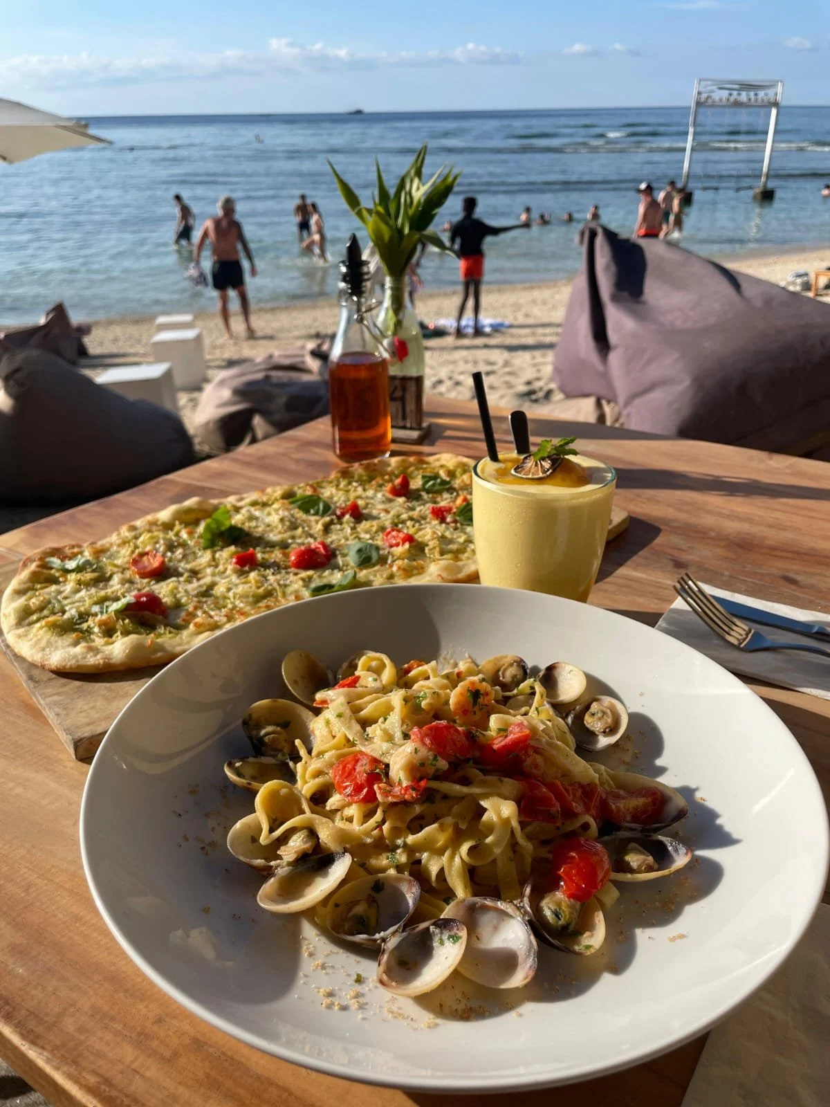 Seafood pasta with clams, cherry tomatoes, and herbs served on a white plate at an outdoor beach restaurant, with a pizza, a cocktail, and a planter in the background, overlooking the beach and ocean with people swimming and playing.