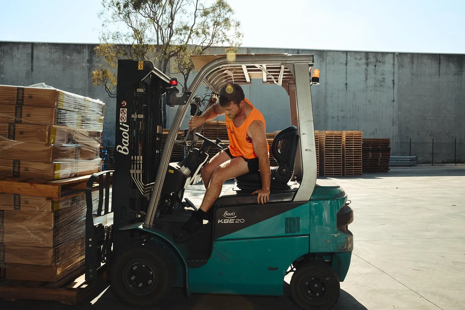 A man wearing an orange sleeveless shirt and black shorts operating a teal forklift in an outdoor warehouse, lifting a pallet of boxed goods.