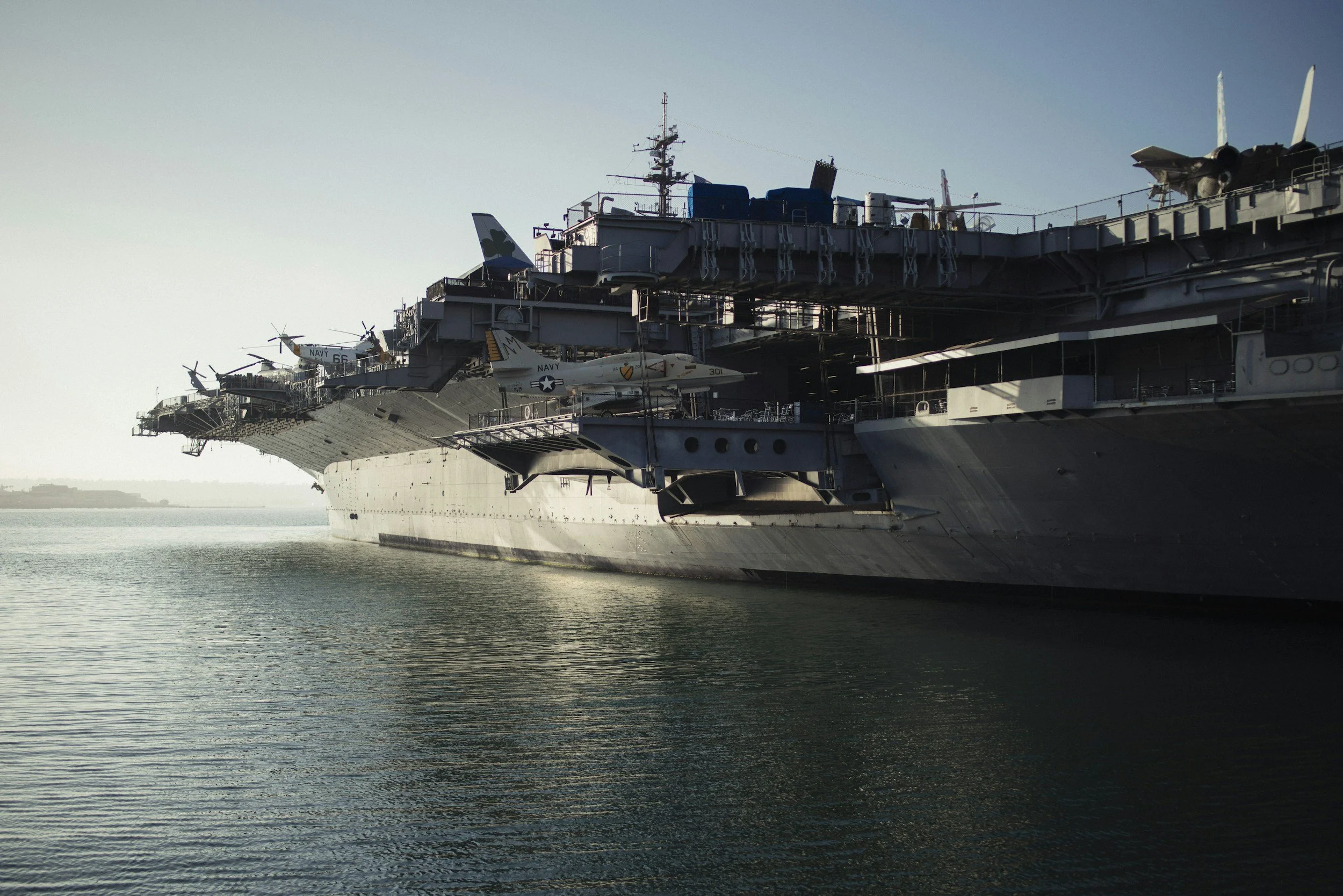 A large military aircraft carrier docked on the water with fighter jets on its deck, against a clear sky.