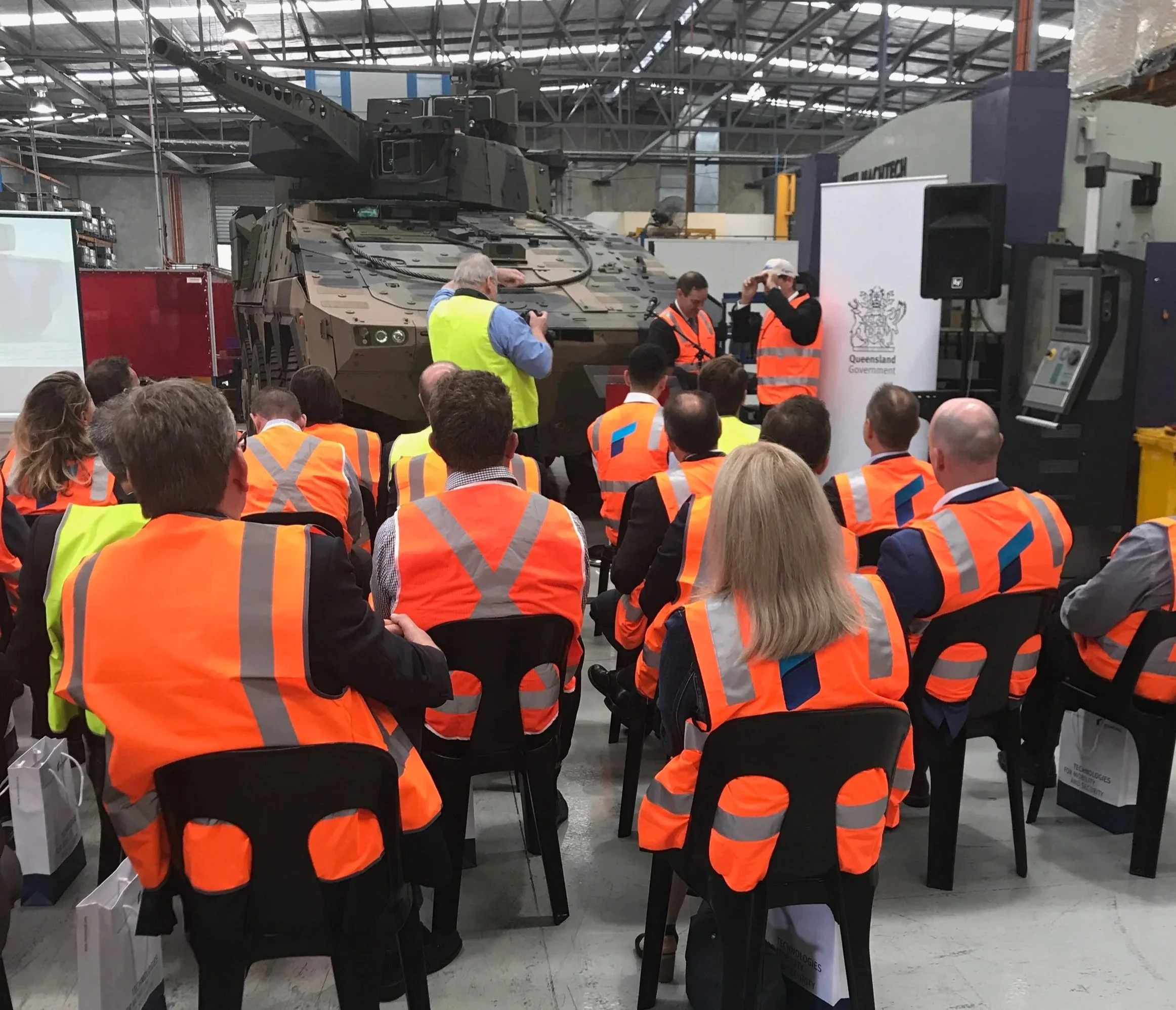 Group of people in orange safety vests attending a presentation about a military tank in a warehouse or factory setting with equipment and machinery.