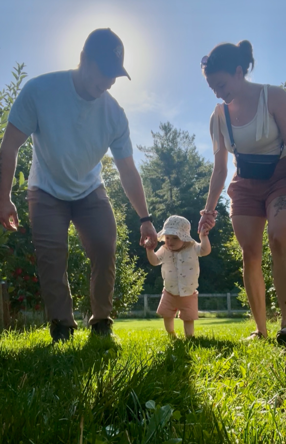 A family of three holding hands on a grassy field with trees, during sunny weather. An adult man and woman are guiding a young child, who is wearing a sun hat, from falling.