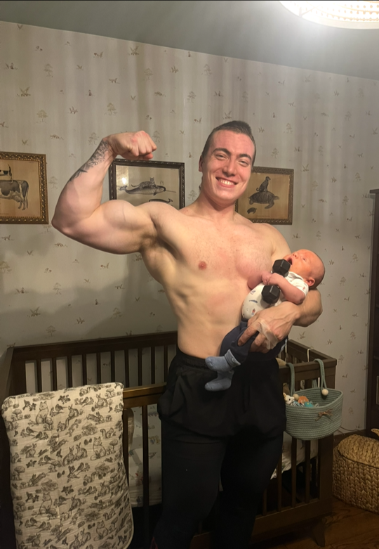 A shirtless man flexing his arm and holding a baby in a room with light-colored wallpaper and framed animal pictures on the wall.