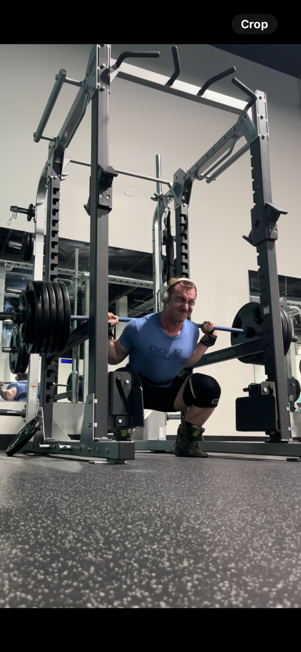 A man performing a barbell back squat in a gym, wearing headphones, a blue T-shirt, knee sleeves, and exercise gloves, in front of a squat rack with weight plates.