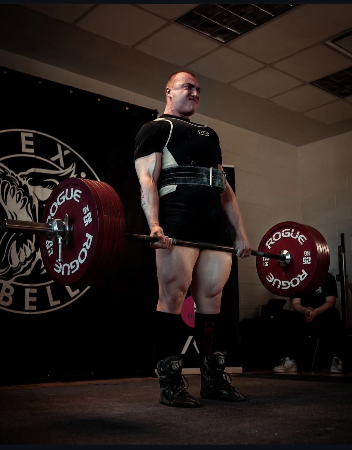 A man in a black and white weightlifting suit is performing a deadlift with a loaded barbell, his face showing strain, in a gym setting.