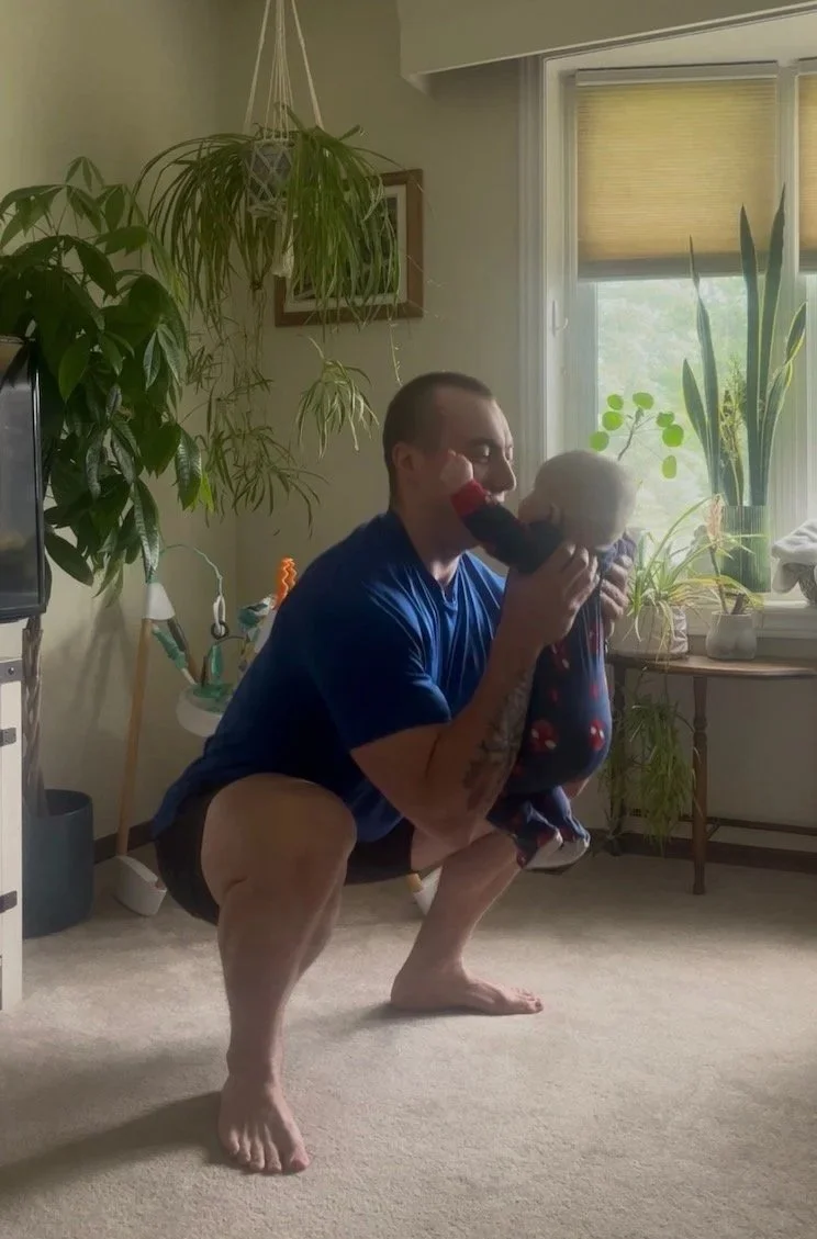 Man crouching on carpeted floor holding a doll near his face in a living room, surrounded by potted plants, with windows and sunlight in the background.