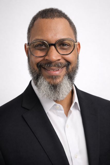 Portrait of owner partner Bernard Barnes, smiling in a black blazer and a white shirt.
