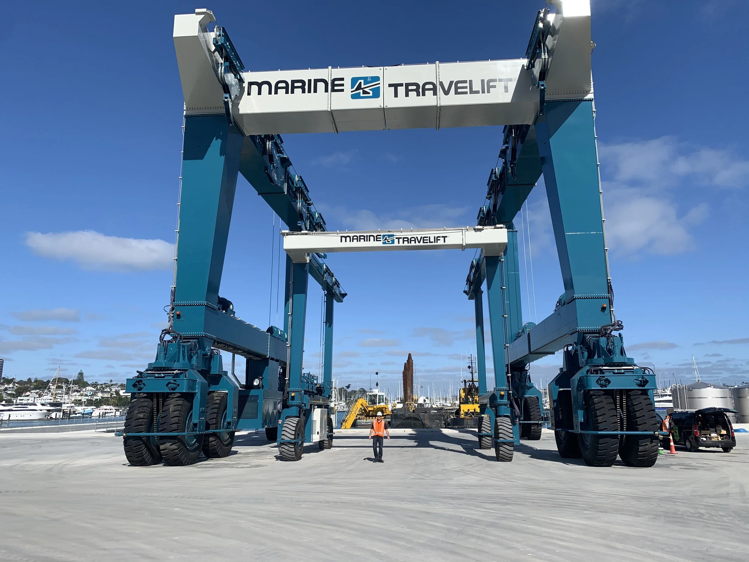Large blue industrial gantry crane with the words marine travelift on it, at a marina or port, under a partly cloudy sky.