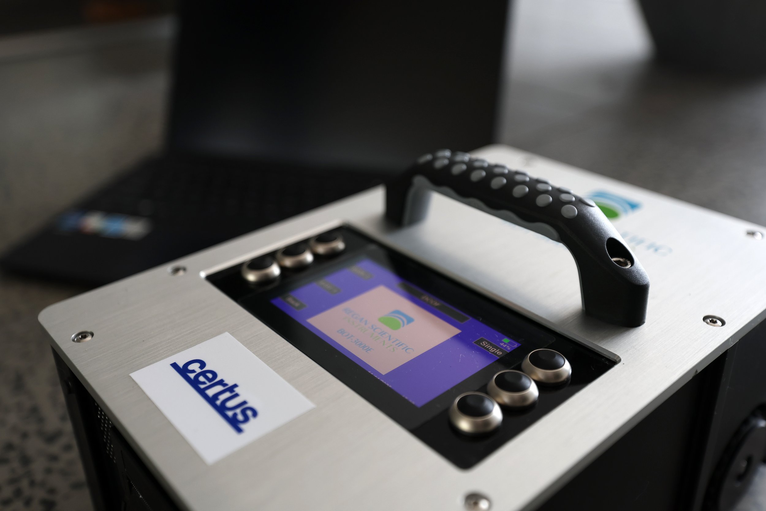 A portable electronic voting or polling device with a touchscreen and buttons, labeled 'Gerritsen', resting on a table with a laptop in the background.