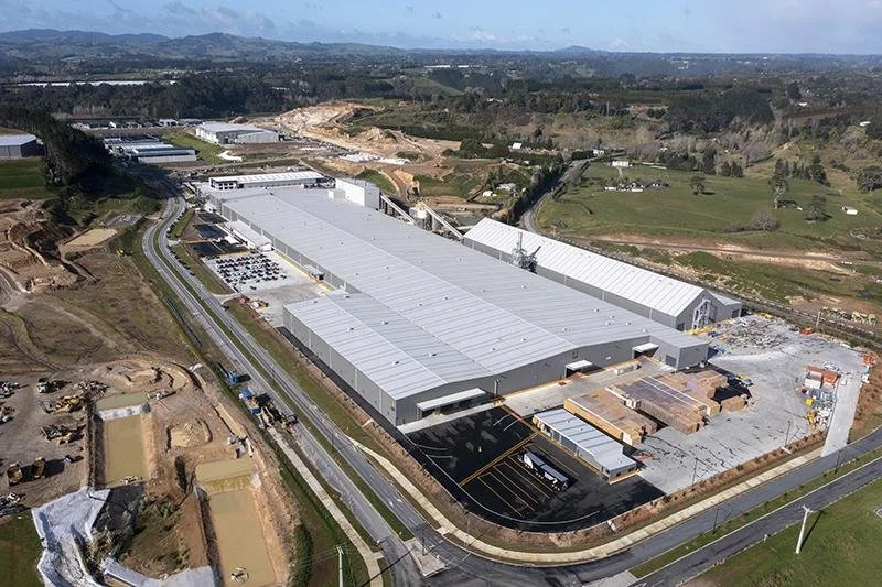 A large industrial warehouse under construction with construction materials around and a parking lot nearby, situated in a rural area with hills and open land.