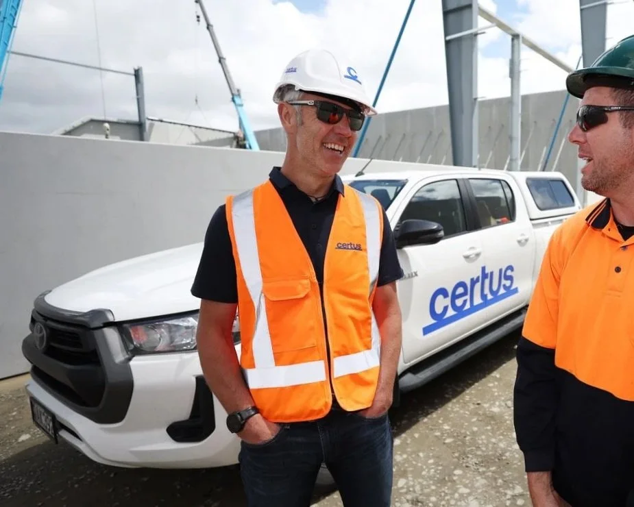 Two men wearing safety vests and helmets standing near a white vehicle with 'certus' written on it, at an outdoor construction or industrial site.