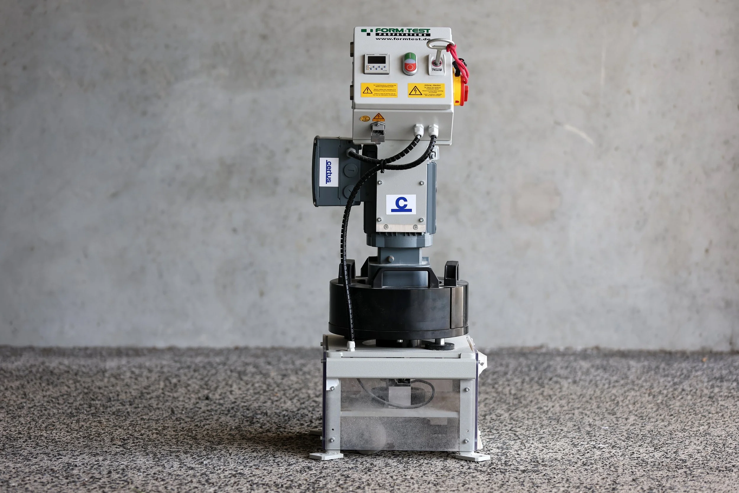 A robotic testing system with a control panel on top, mounted on a cylindrical black base, placed on a table with speckled gray surface against a plain wall background.