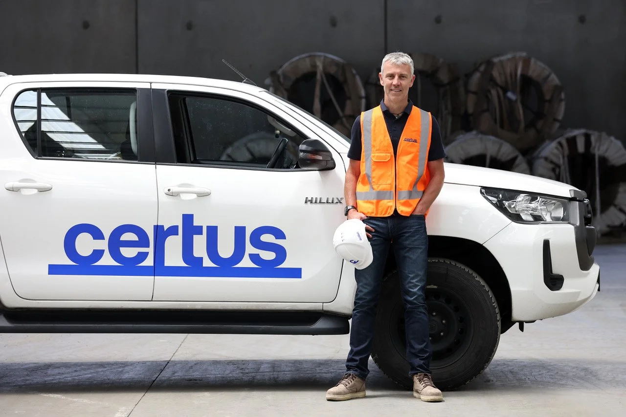 A man in a black shirt, jeans, and tan work boots standing next to a white vehicle with 'certus' written in blue on the side, holding a white safety helmet and wearing an orange safety vest, in an industrial setting.