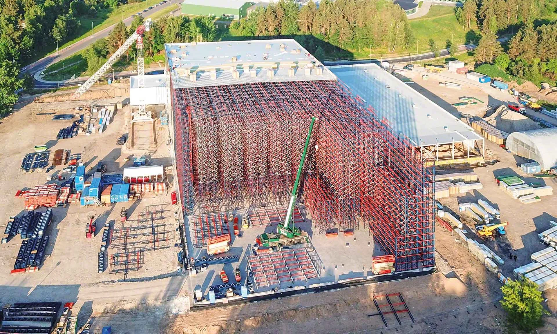 An aerial view of a construction site with a building framework, a crane, construction materials, and surrounding greenery.