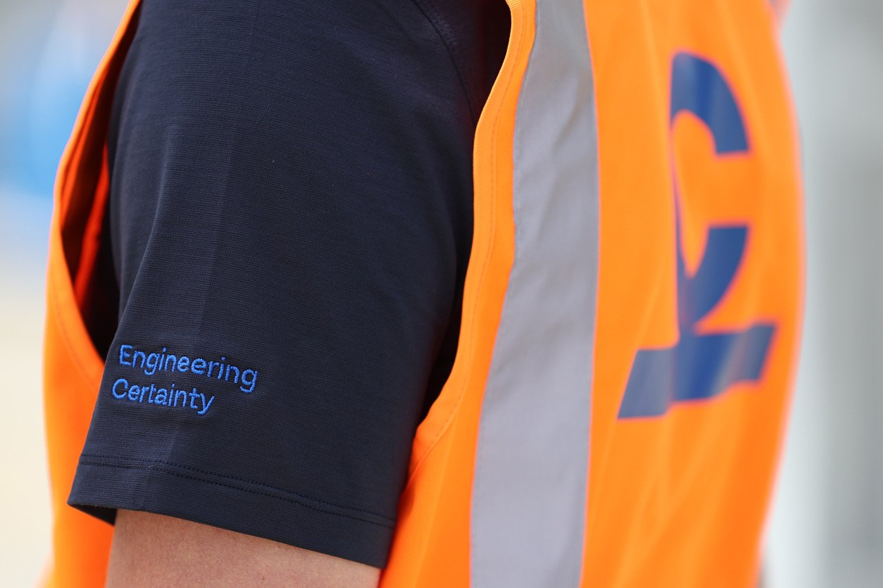 Close-up of a person's shoulder wearing a dark shirt with the words 'Engineering Certainty' embroidered in blue, and an orange safety vest with a large blue logo.