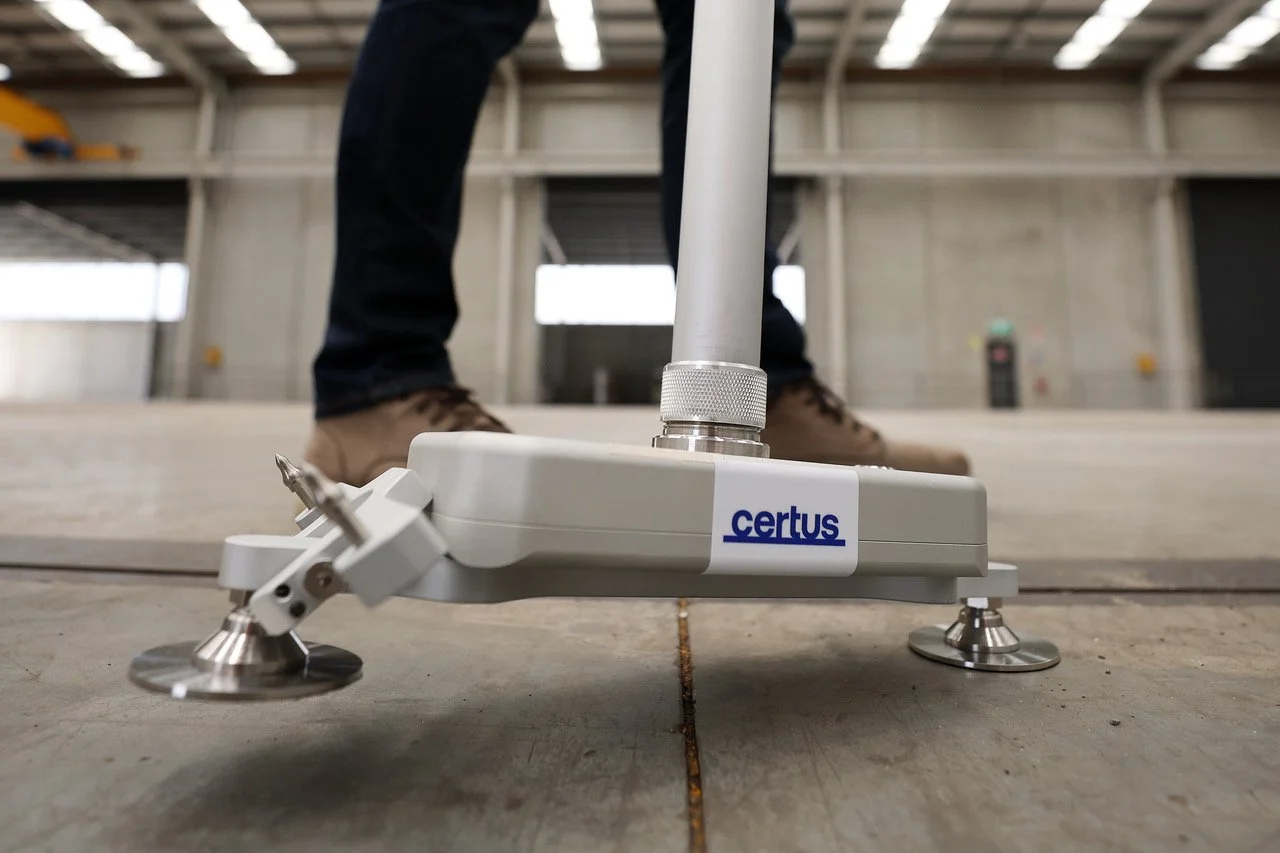 Close-up of a Certus laser alignment tool on concrete floor, used for precision measurements in construction, with legs and part of a person's legs and shoes in the background.