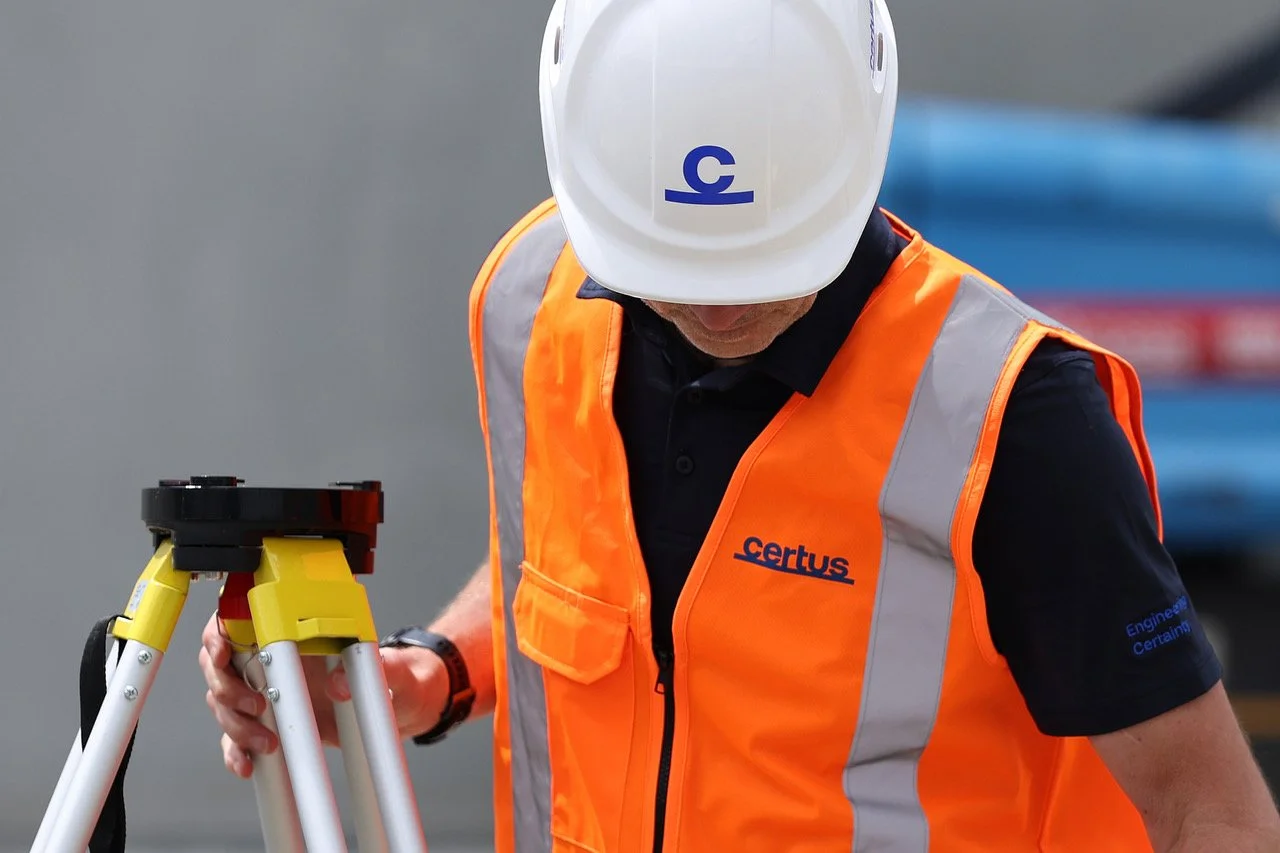 A construction worker wearing an orange safety vest and white hard hat, using surveying equipment.