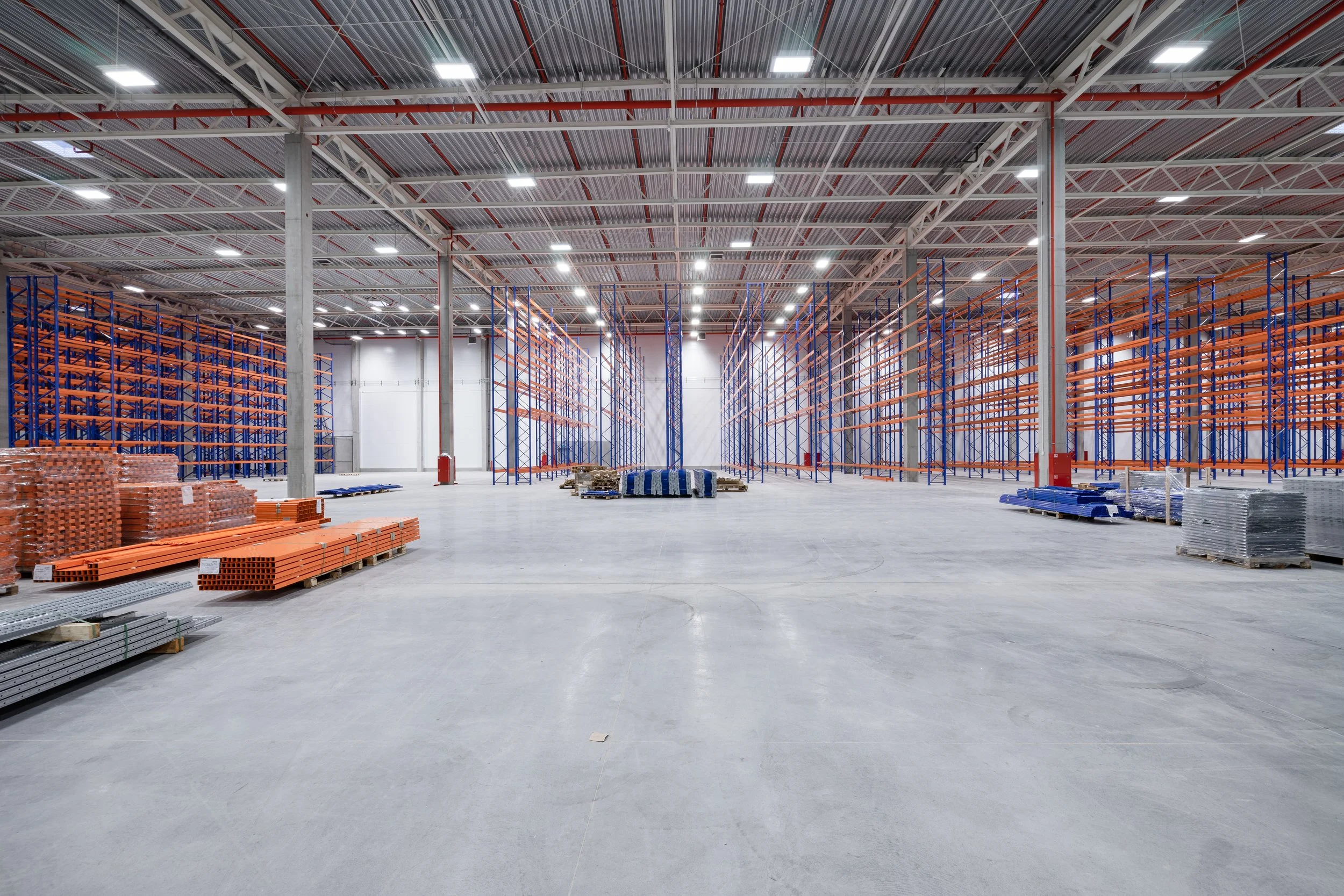 Empty warehouse with industrial shelving units, construction materials, and equipment, under a high metal ceiling with lighting fixtures.